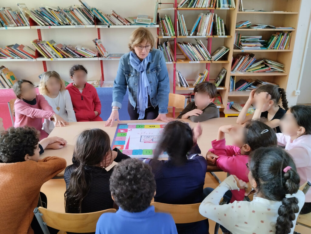 Photo d'une sensibilisation : nous voyons un membre de CAP'école devant une classe rassemblée autour d'une table et de notre jeu de plateau.