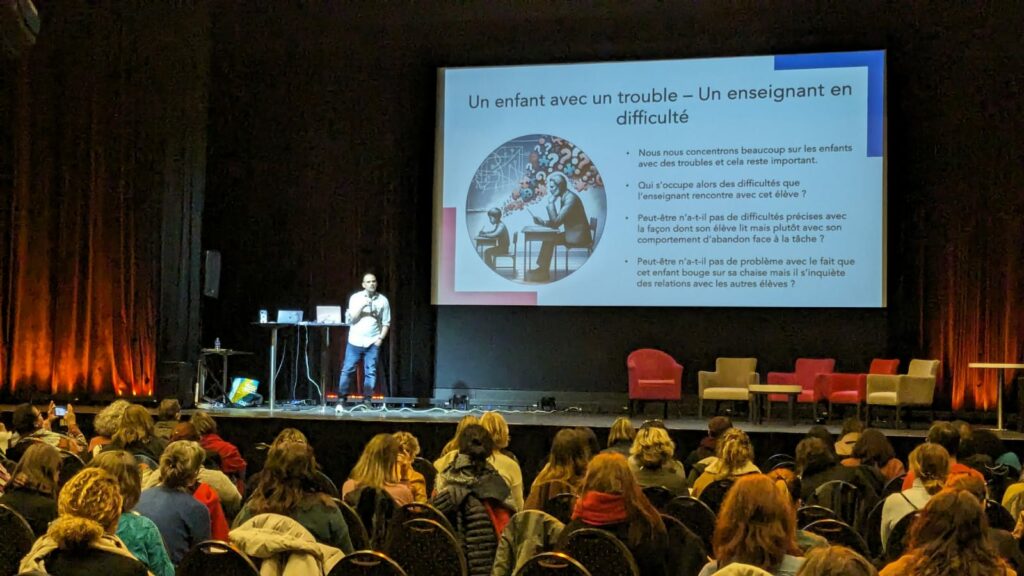 Photo de Sébastien Henrard lors du colloque 2023. On le voit sur scène avec le diaporama sur lequel est écrit : un enfant avec un trouble, un enseignant en difficulté.